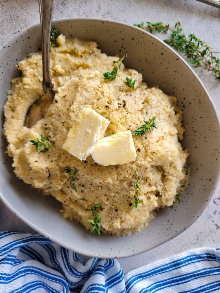 cauliflower mashed potatoes in a bowl topped with butter and fresh thyme, more thyme in the background, spoon in the bowl