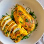 two yellow chicken breasts on a bed of arugula in a white bowl garnished with fresh chopped parsley.