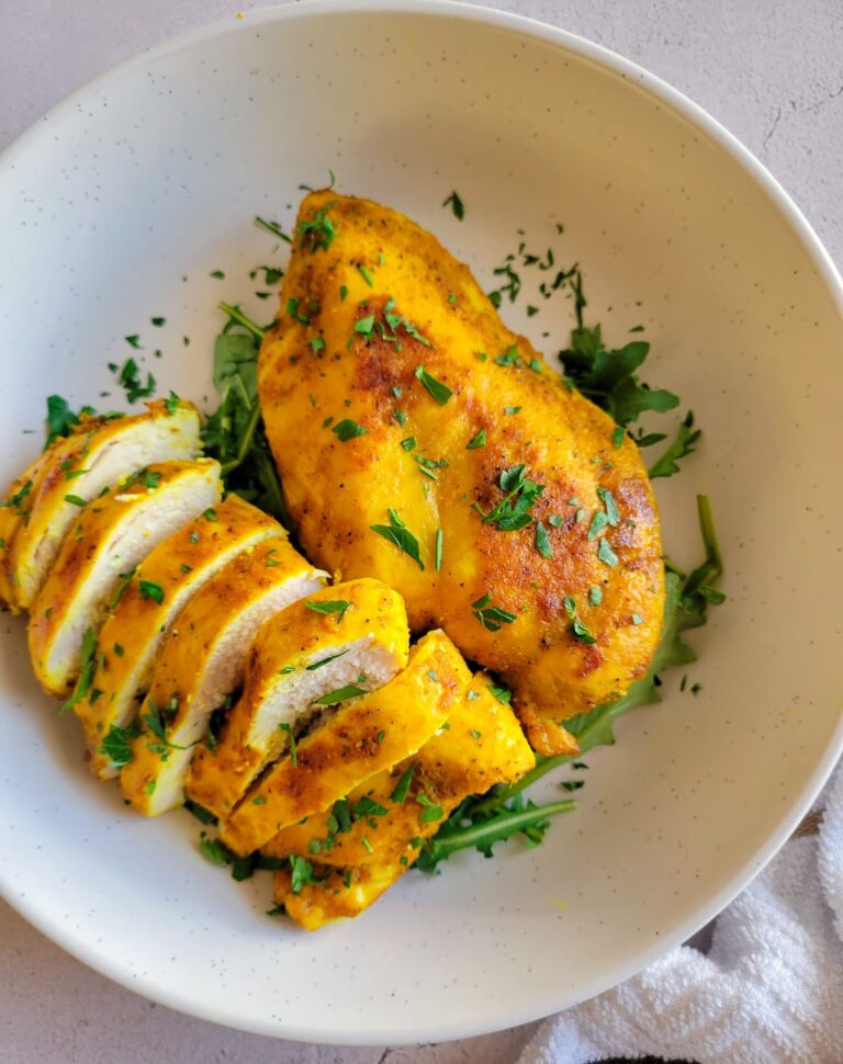 two yellow chicken breasts on a bed of arugula in a white bowl garnished with fresh chopped parsley.