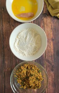 3 bowls - one of beaten egg, one of flour and one of a quinoa coating mixture, chicken tender in the bowl of egg