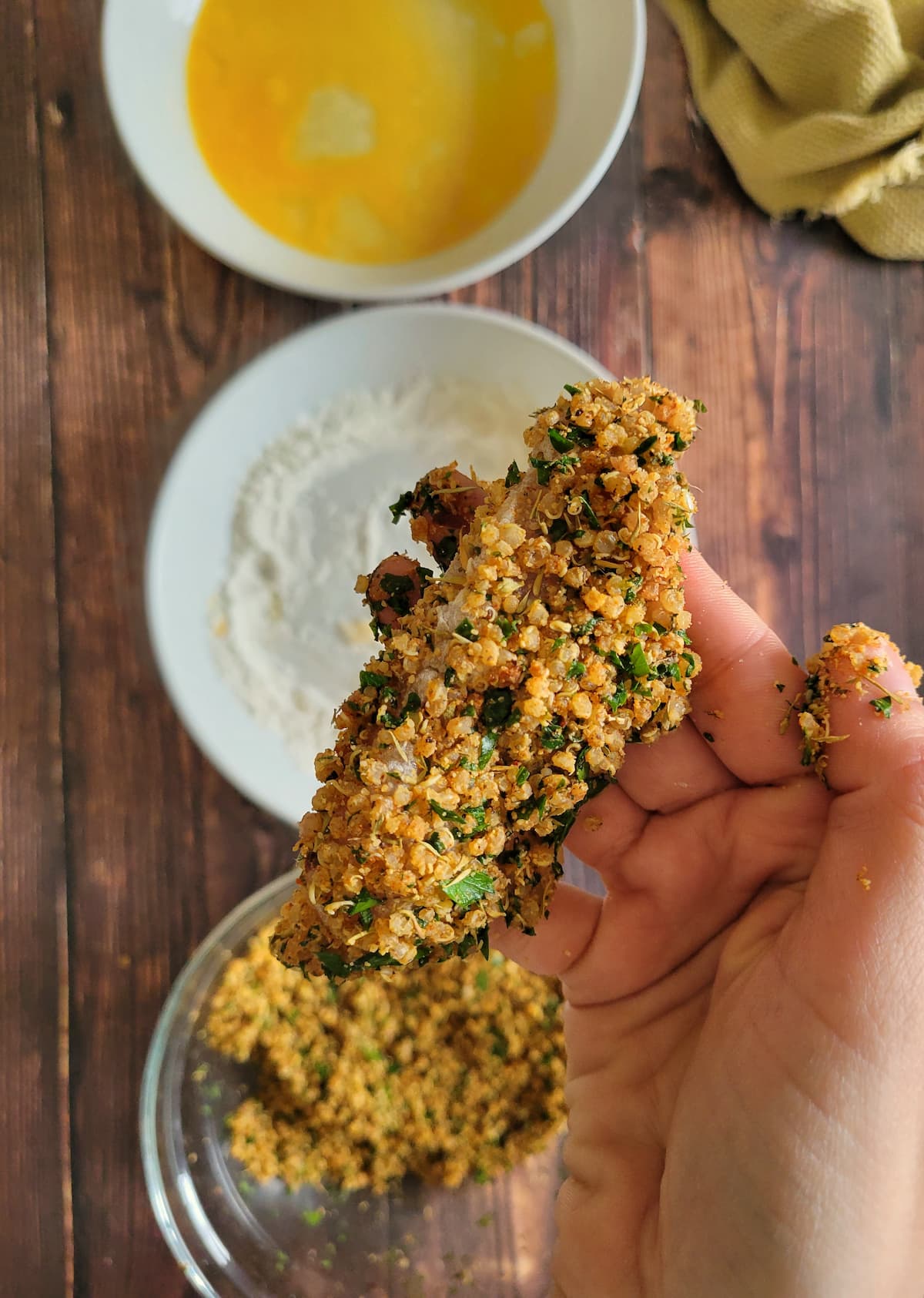 hand holding a quinoa chicken tender over two bowls of egg and flour