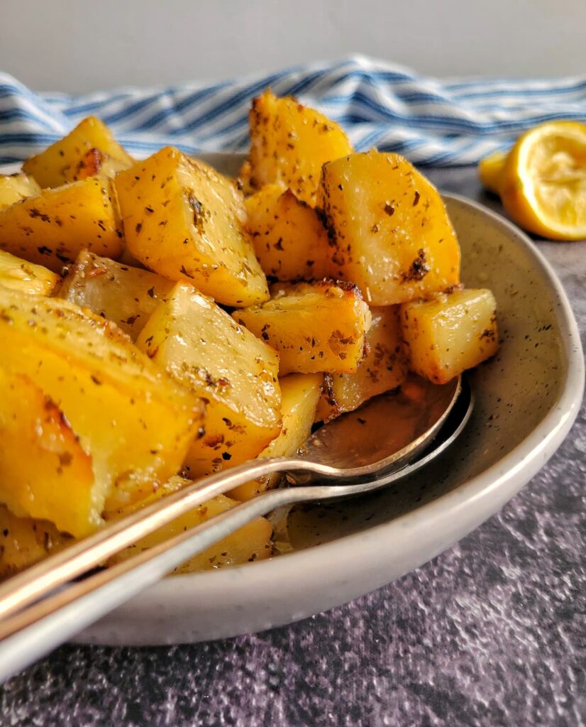 side view of greek roasted potatoes in a bowl with two spoons, halved lemons in the background