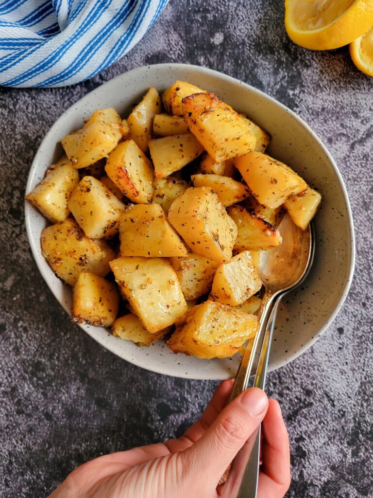 roasted and seasoned potatoes in a bowl with two spoons, hand on the spoons, lemon halves in the background
