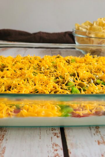 side view of layered nacho dip topped with shredded cheddar cheese and sliced jalapenos, bowl of tortilla chips in the background