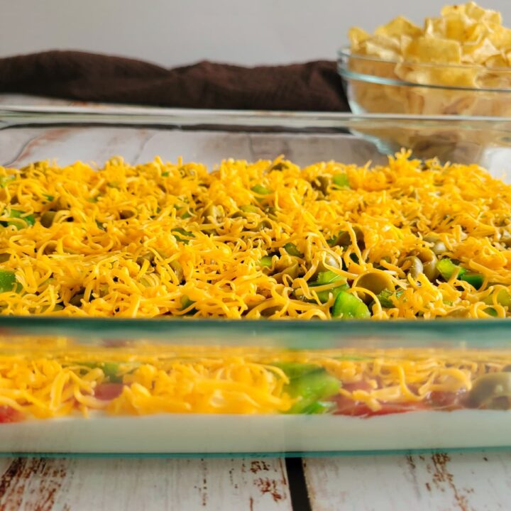 side view of layered nacho dip topped with shredded cheddar cheese and sliced jalapenos, bowl of tortilla chips in the background
