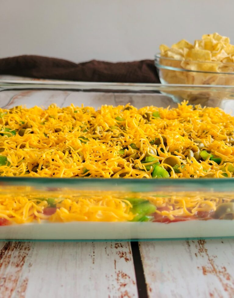 side view of layered nacho dip topped with shredded cheddar cheese and sliced jalapenos, bowl of tortilla chips in the background