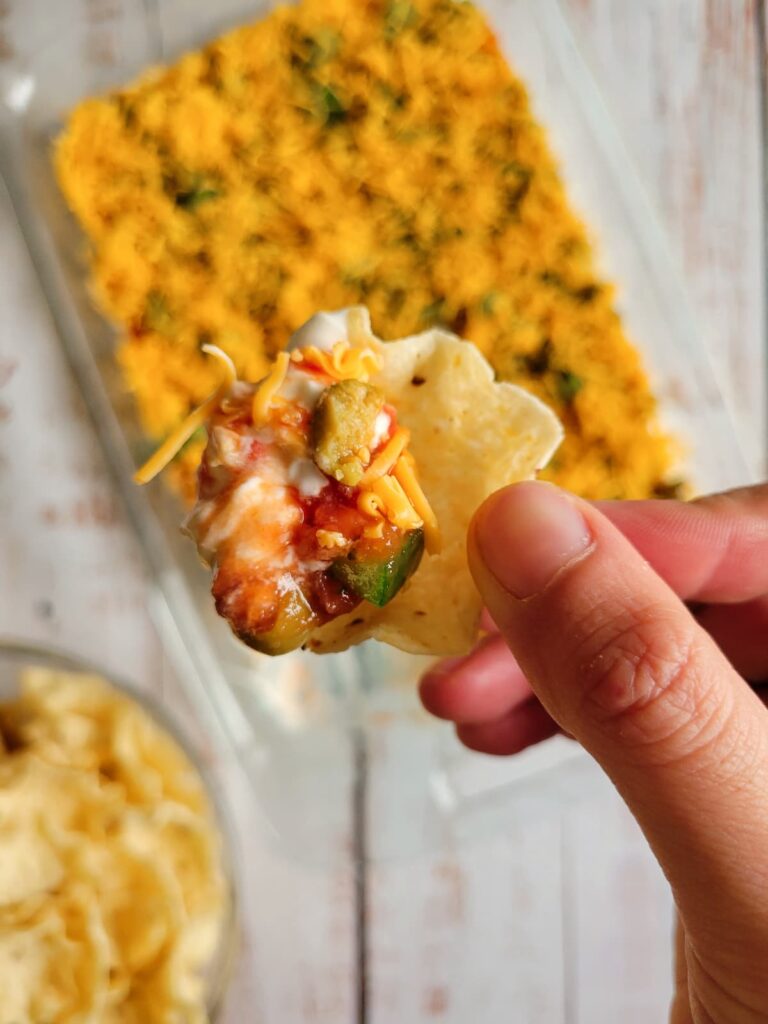 hand holding a tortilla chip with nacho dip on it over a dish with the rest of it