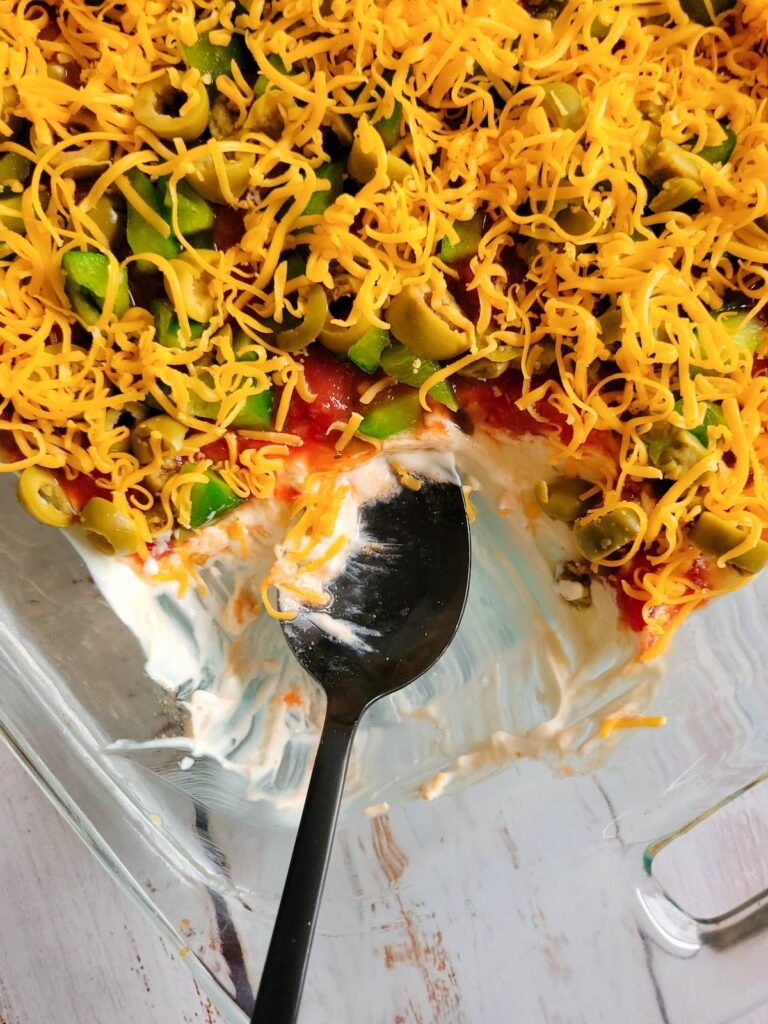 spoon in a dish with layered nacho dip with sour cream, salsa, cheddar, green pepper and green olives
