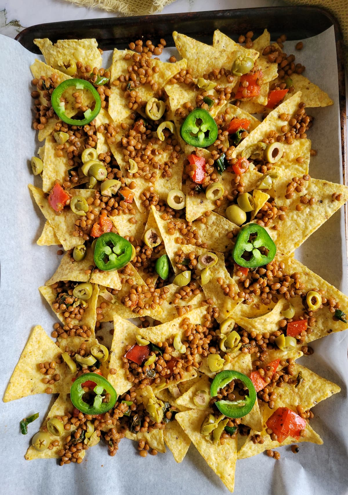 lentils, jalapenos, red peppers and green olives piled on top of tortilla chips on a parchment lined baking sheet
