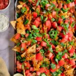 tortilla chips on a baking sheet loaded with chopped tomatoes, green onions, lentils, green olives, cheddar cheese, tomatoes, sour cream and salsa on the side
