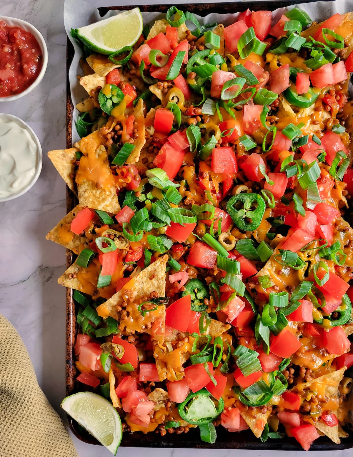 tortilla chips on a baking sheet loaded with chopped tomatoes, green onions, lentils, green olives, cheddar cheese, tomatoes, sour cream and salsa on the side