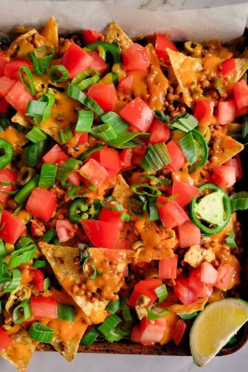 half view of tortilla chips on a baking sheet loaded with chopped tomatoes, green onions, lentils, green olives and cheddar cheese, lime wedge