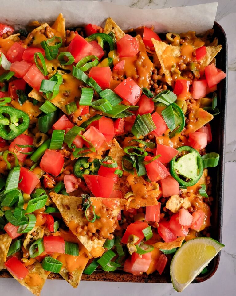 half view of tortilla chips on a baking sheet loaded with chopped tomatoes, green onions, lentils, green olives and cheddar cheese, lime wedge