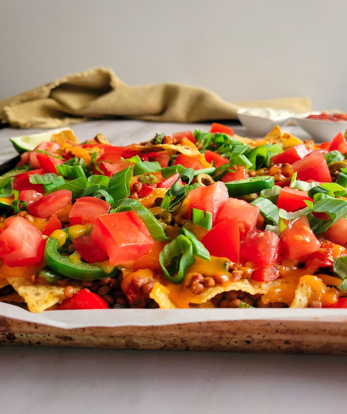side view of lentil and veggie nachos on parchment lined baking sheet, sour cream and salsa in the background