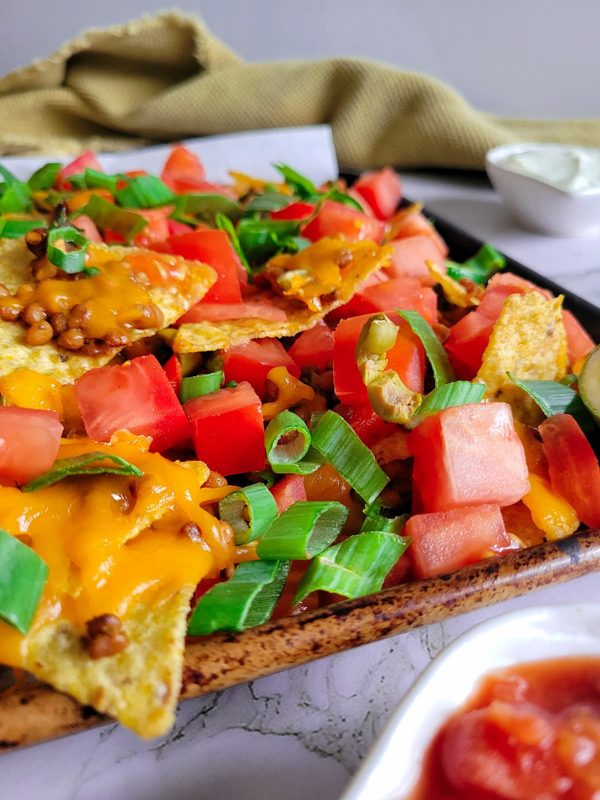 side view of tortilla chips on a baking sheet loaded with chopped tomatoes, green onions, lentils, green olives and cheddar cheese