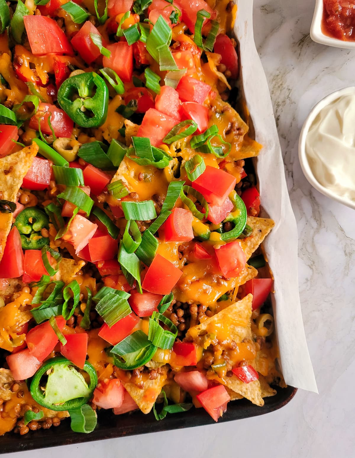 loaded lentil nachos on a parchment lined baking sheet, sour cream and salsa on the side
