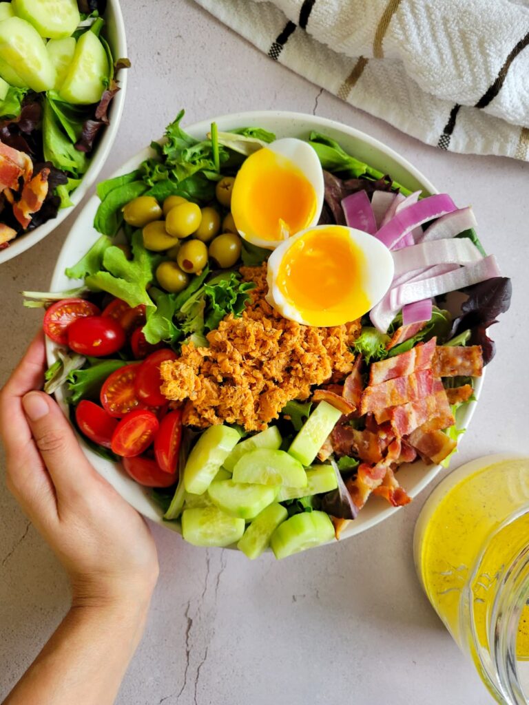 hand holding a bowl of cobb salad with canned salmon, soft boiled eggs, cucumbers, cherry tomatoes, bacon, red onions and olives, dressing and another bowl of salad in the background