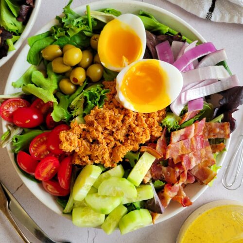 salmon cobb salad with soft boiled eggs, cucumbers, cherry tomatoes, olives, bacon, red onions and greens, Dressing, forks and another bowl of salad in the background