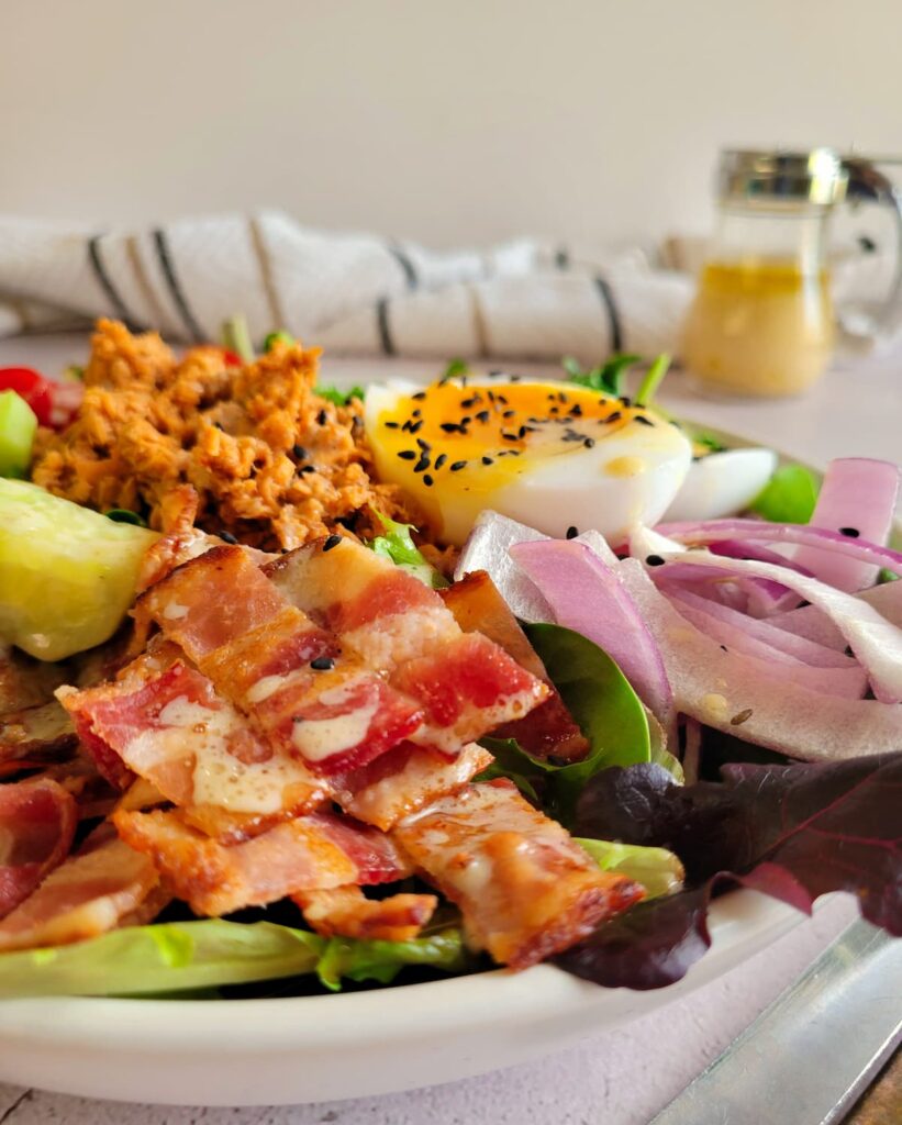 salmon cobb salad with soft boiled eggs, cucumbers, cherry tomatoes, olives, bacon, red onions and greens, jar of dressing in the background