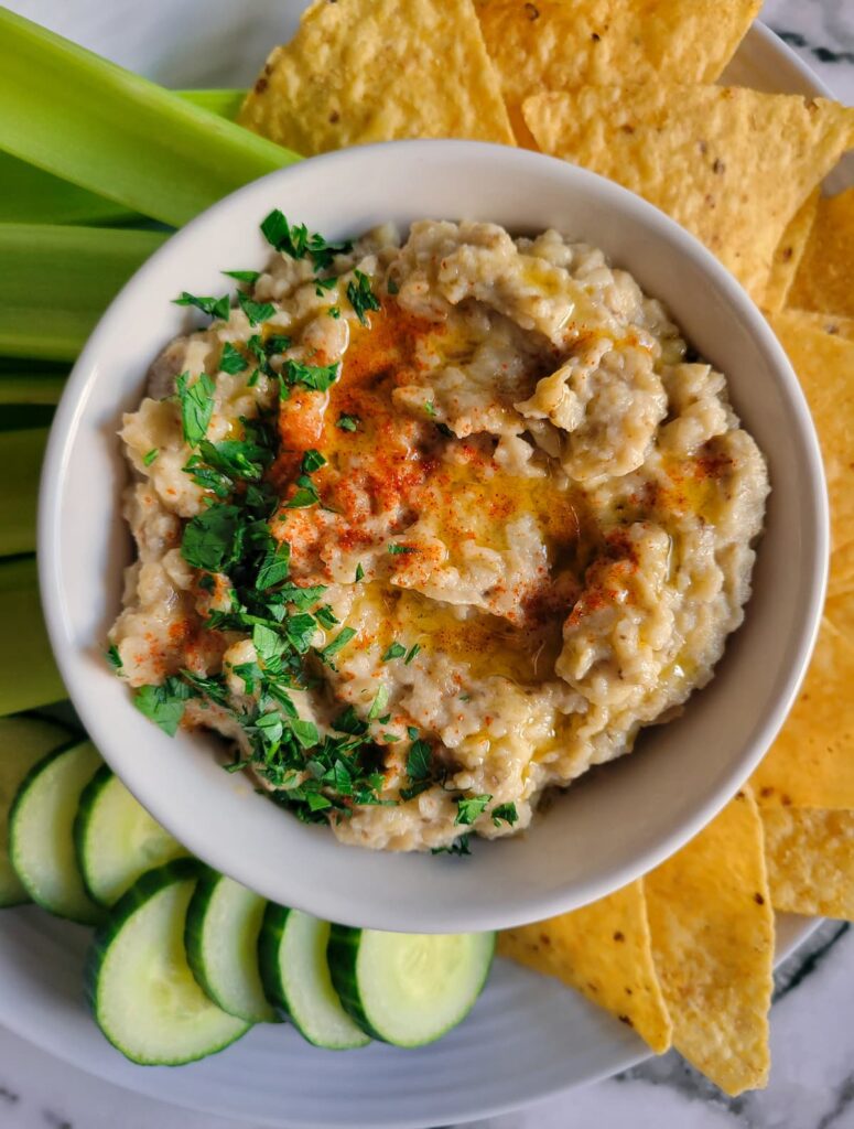 bowl of baba ganoush topped with fresh chopped parsley, olive oil and smoked paprika on a plate with tortilla chips, celery stick and cucumber slices