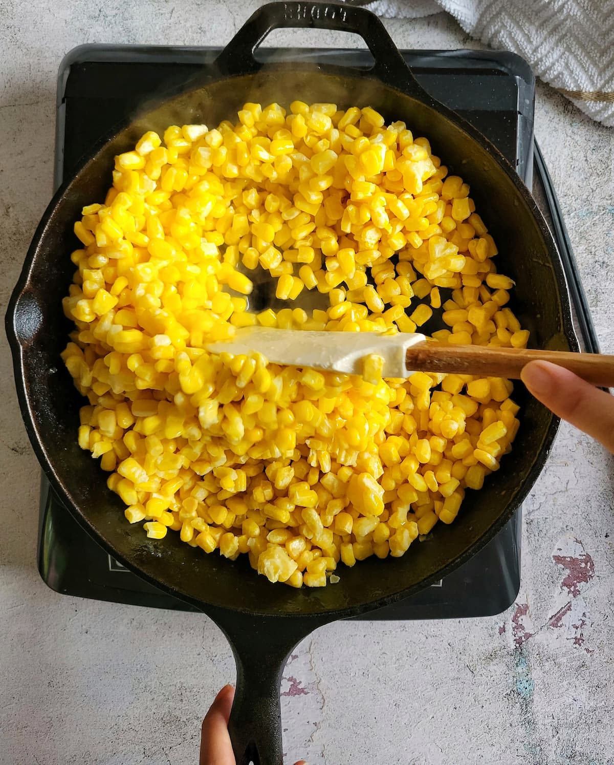 hand with a rubber spatula stirring corn in a skillet on a burner