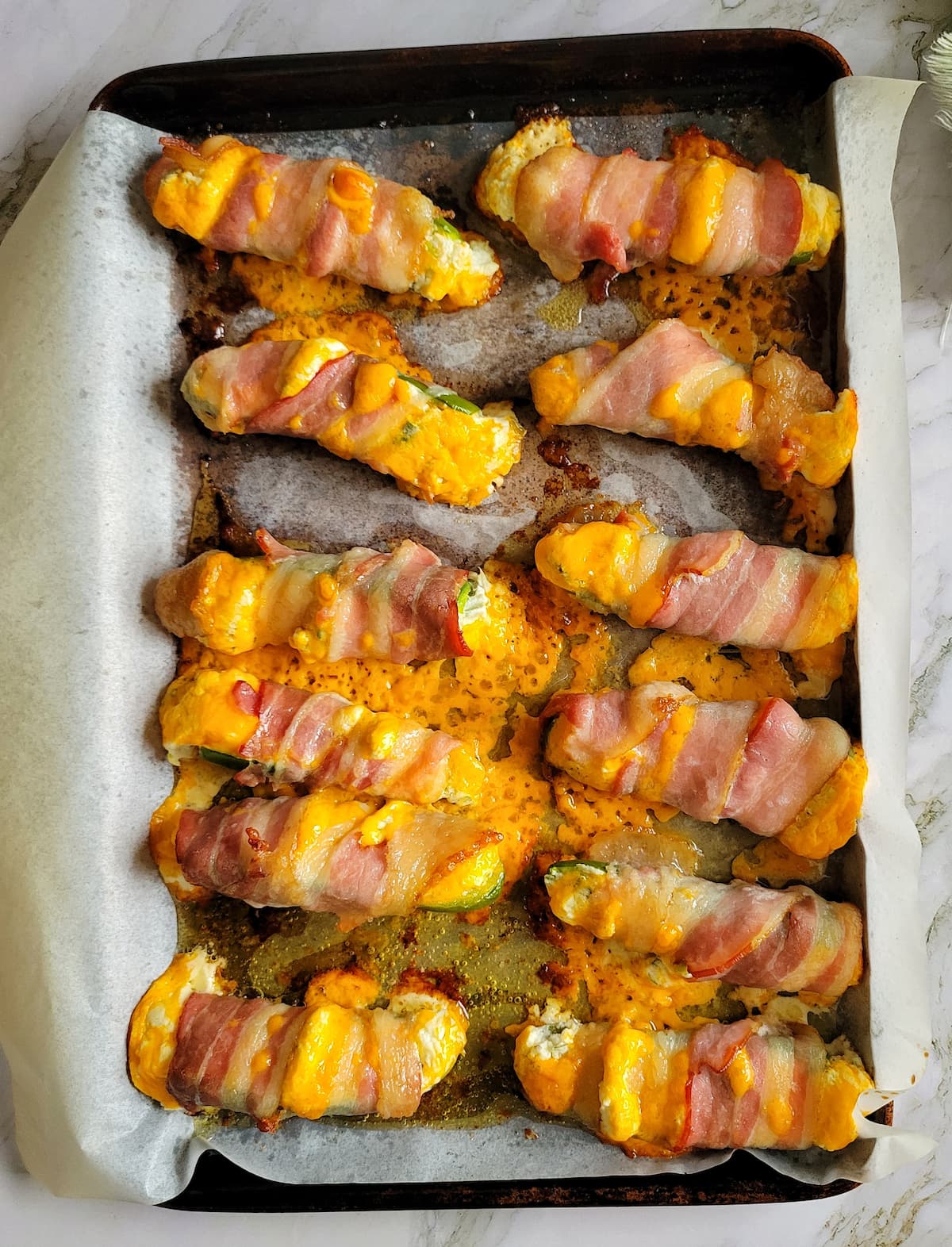 cooked jalapeno poppers wrapped in bacon on a parchment lined baking sheet with lots of cheddar cheese around