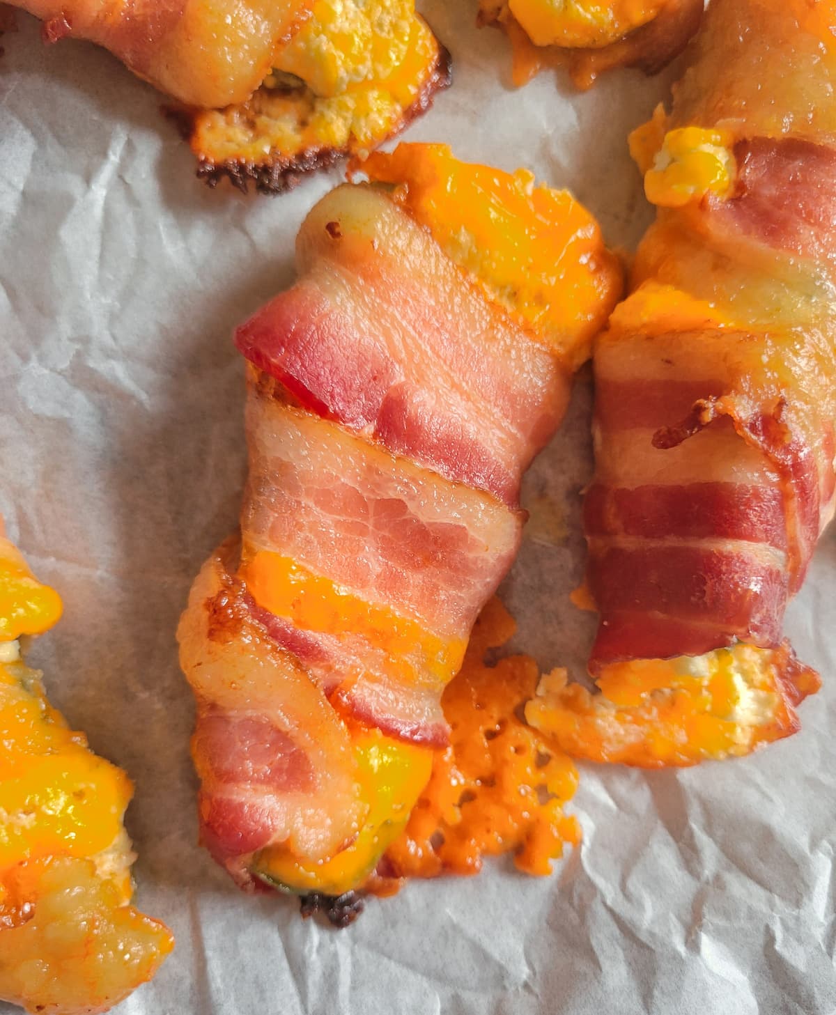 close up of a jalapeno popper wrapped in bacon with cheddar cheese on a parchment lined baking sheet with the rest of them