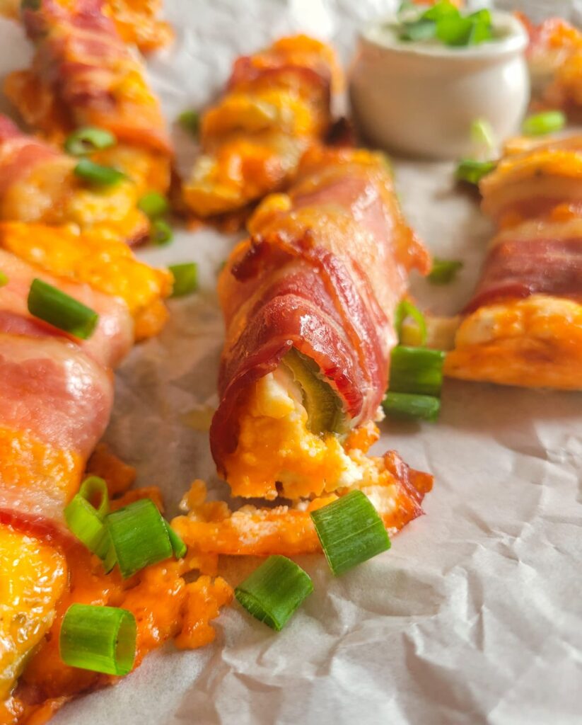 close up of a jalapeno popper wrapped in bacon with cheddar cheese and green onions on a parchment lined baking sheet with the rest of them,