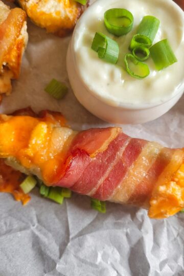 close up of a jalapeno popper wrapped in bacon with cheddar cheese and green onions on a parchment lined baking sheet with the rest of them, white dipping sauce with chopped green onions in a ramekin
