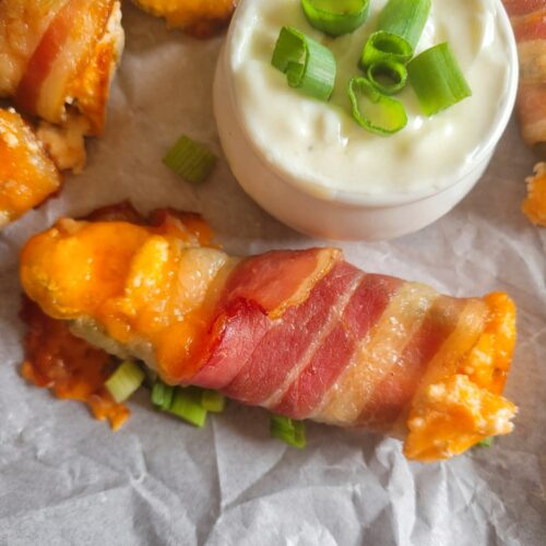 close up of a jalapeno popper wrapped in bacon with cheddar cheese and green onions on a parchment lined baking sheet with the rest of them, white dipping sauce with chopped green onions in a ramekin