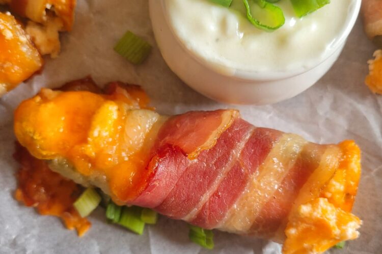 close up of a jalapeno popper wrapped in bacon with cheddar cheese and green onions on a parchment lined baking sheet with the rest of them, white dipping sauce with chopped green onions in a ramekin