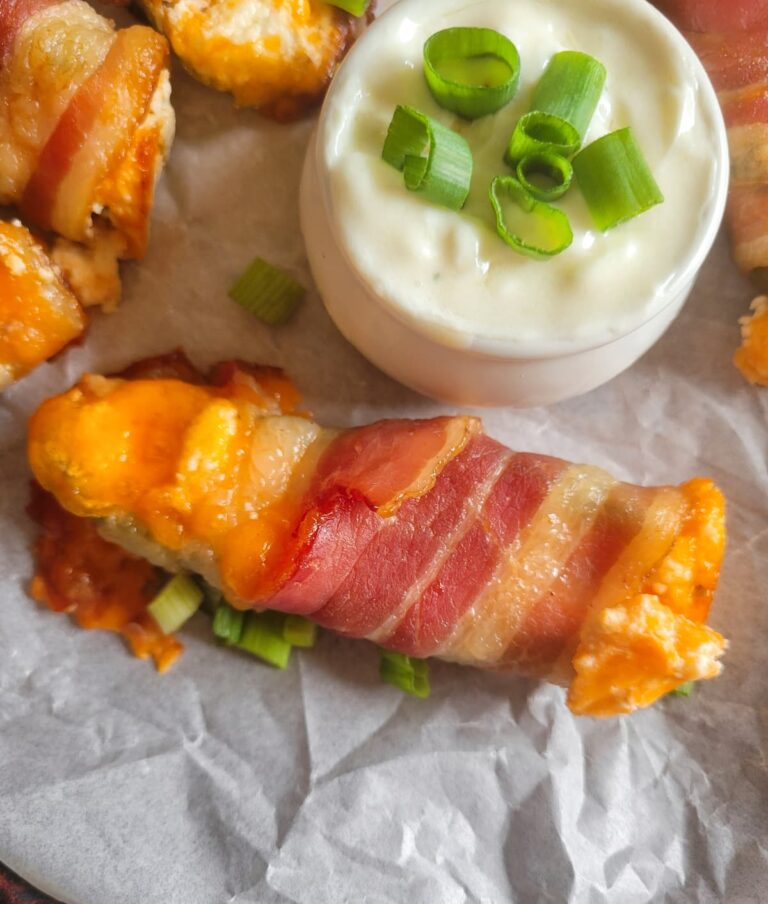 close up of a jalapeno popper wrapped in bacon with cheddar cheese and green onions on a parchment lined baking sheet with the rest of them, white dipping sauce with chopped green onions in a ramekin