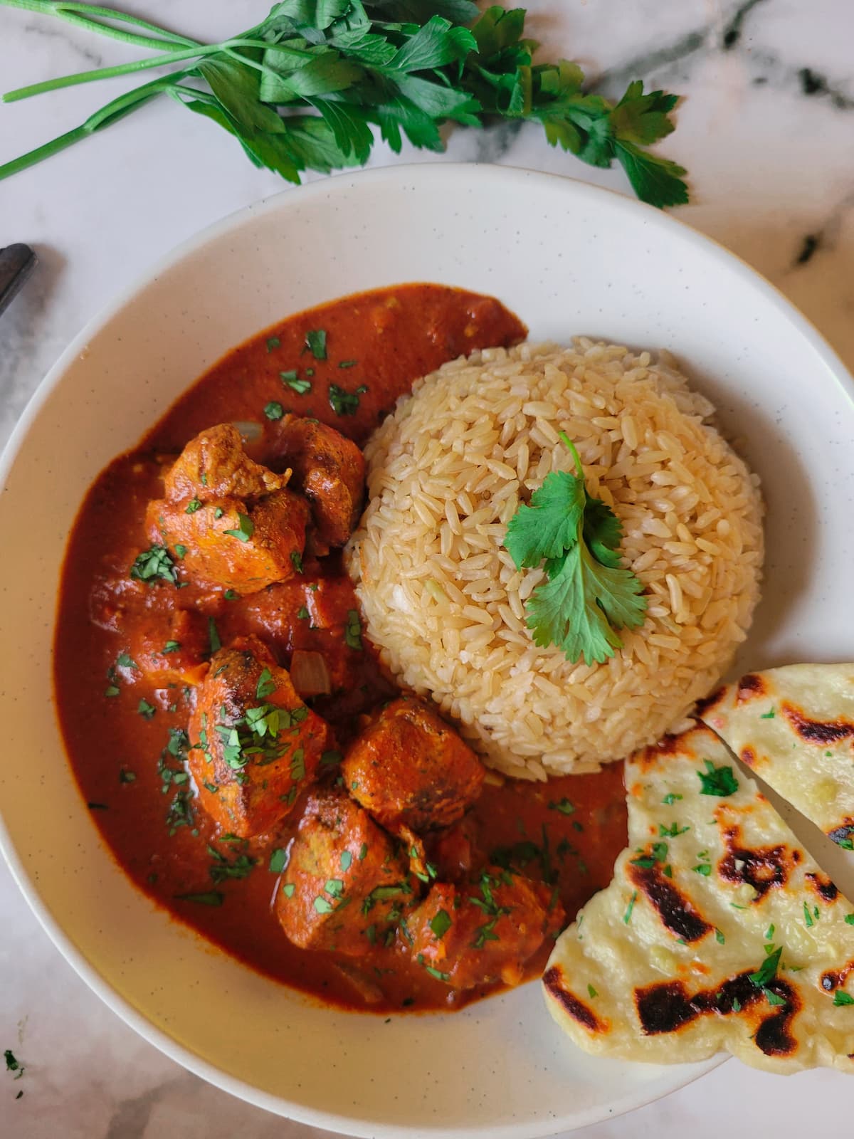 bowl of butter chicken served with rice and naan bread, garnished with fresh chopped parsley