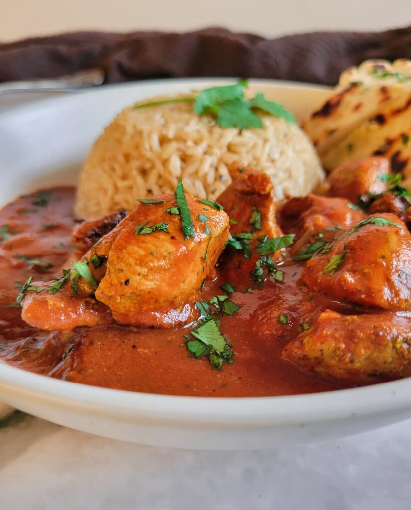 side view of a bowl of butter chicken served with naan and rice, garnished with chopped cilantro