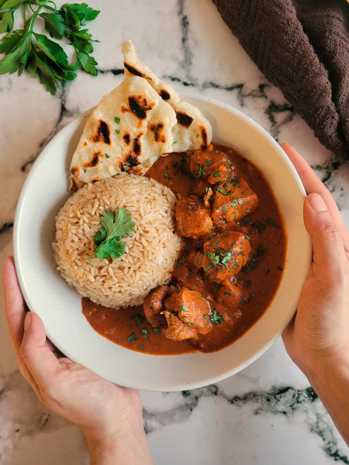 hands holding a bowl of butter chicken served with rice and naan bread, garnished with fresh chopped parsley