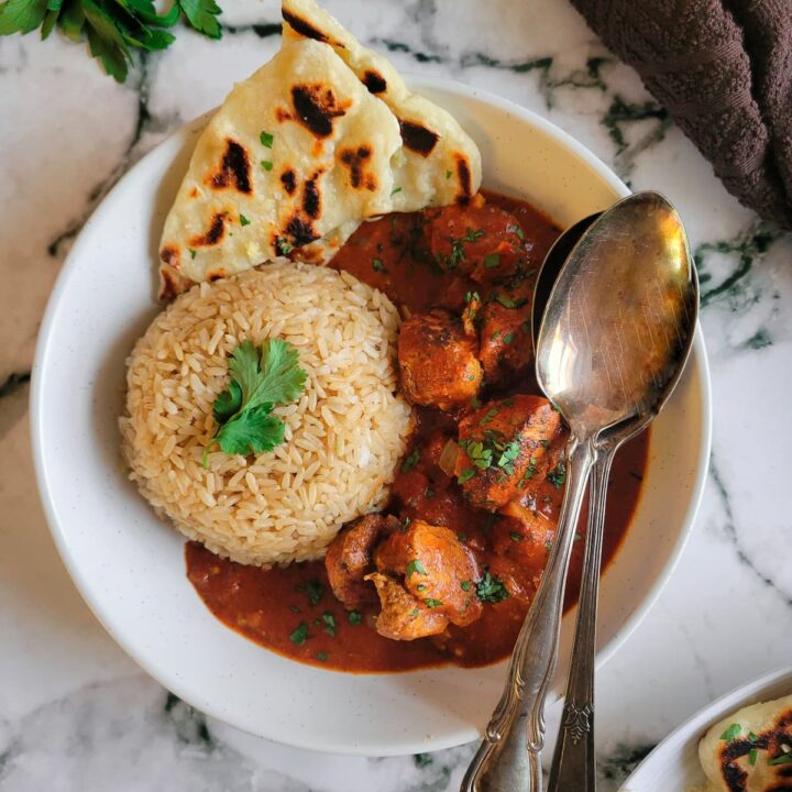 bowl of butter chicken served with rice and naan bread, garnished with fresh chopped parsley, two spoons in the bowl, more naan on the side