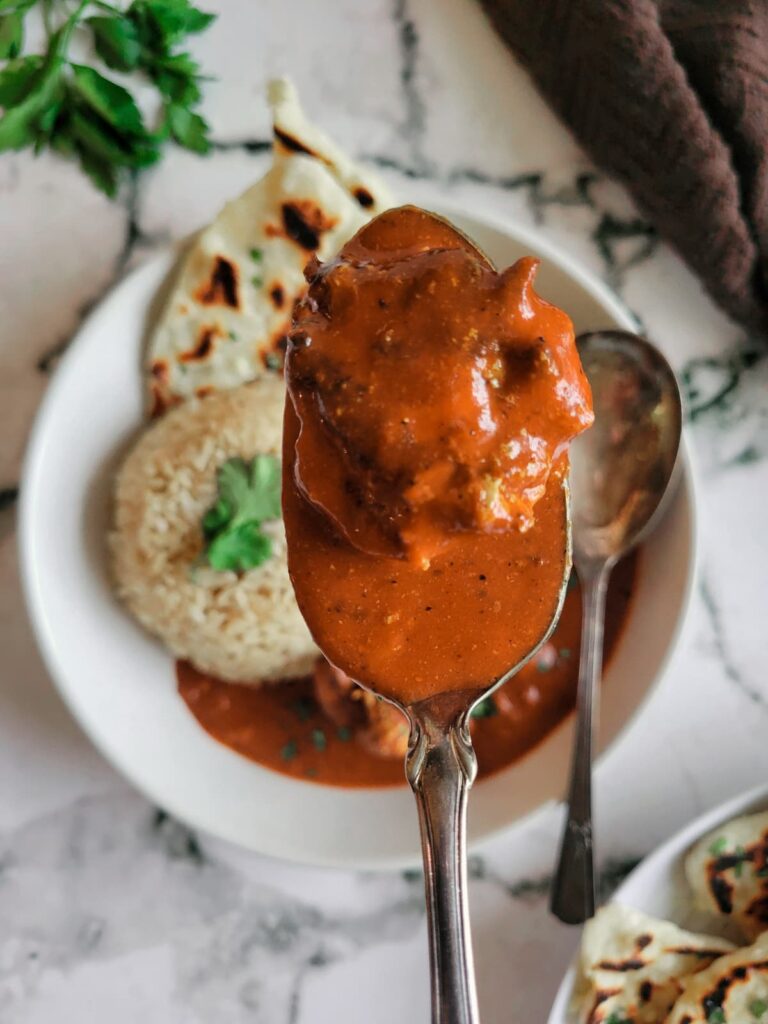 spoonful of butter chicken over a bowl with naan, rice and more butter chicken