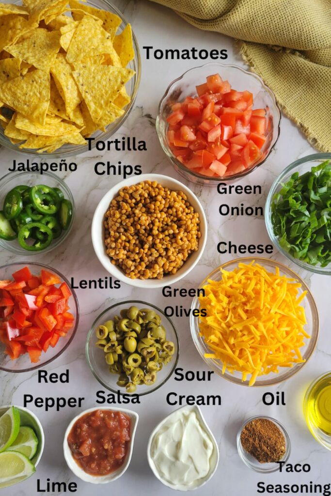 ingredients for lentil nachos - lentils, tomatoes, green onions, jalapeno, red peppers, taco seasoning, oil, sour cream, salsa, lime, green olives, tortilla chips, cheese