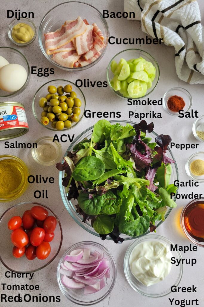 ingredients for salmon cobb salad - salmon, apple cider vinegar, olives, greens, cucumber, bacon, dijon, eggs, smoked paprika, salt, pepper, garlic powder, maple syrup, greek yogurt, red onions, cherry tomatoes, olive oil