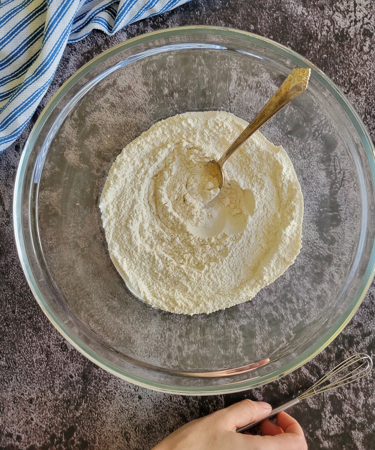 bowl of flour with a spoon in it, hand with a little whisk beside it