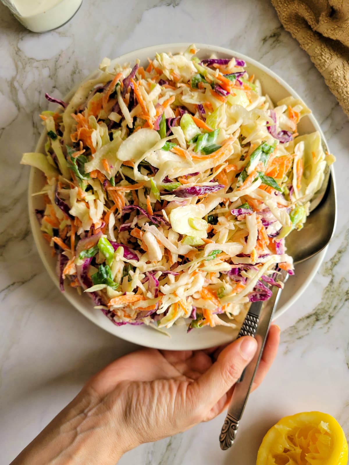 hand holding a bowl of coleslaw with two spoons, lemon on the side