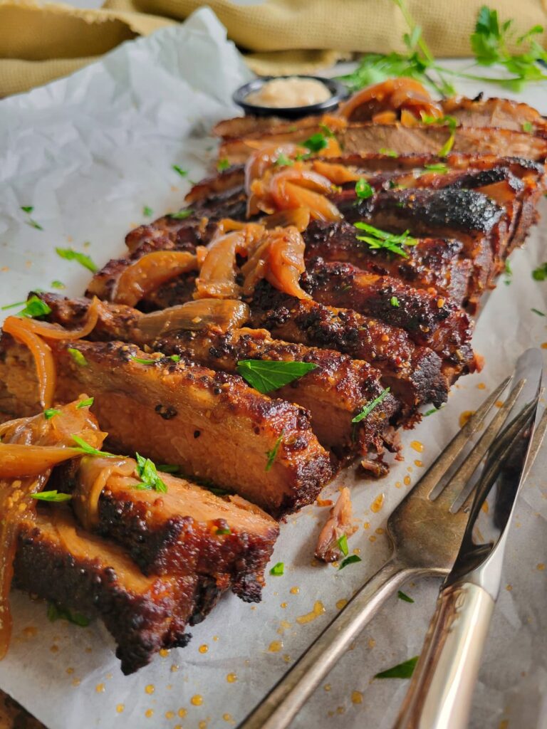 side view of sliced brisket with onions and fresh herbs on a parchment lined baking sheet, sauce and herbs in the background, fork and knife on the tray