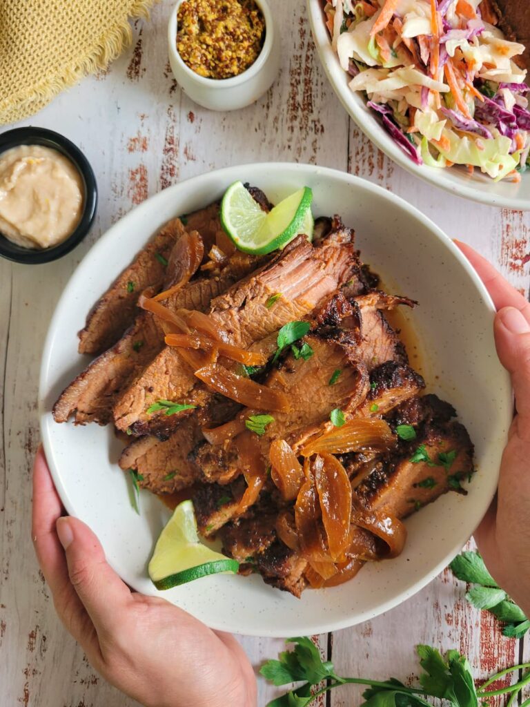 hands holding a bowl of sliced brisket with onions, lime, fresh herbs, sauces and coleslaw in the background