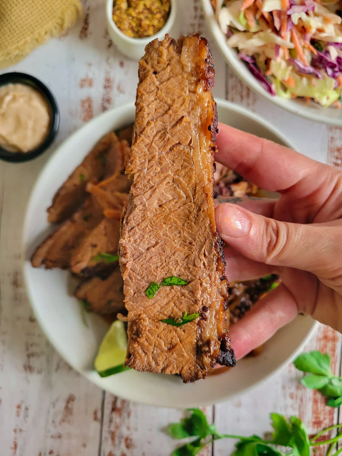 hand holding up a whole piece of brisket over a bowl with the rest, sauces, lime, herbs and coleslaw in the background