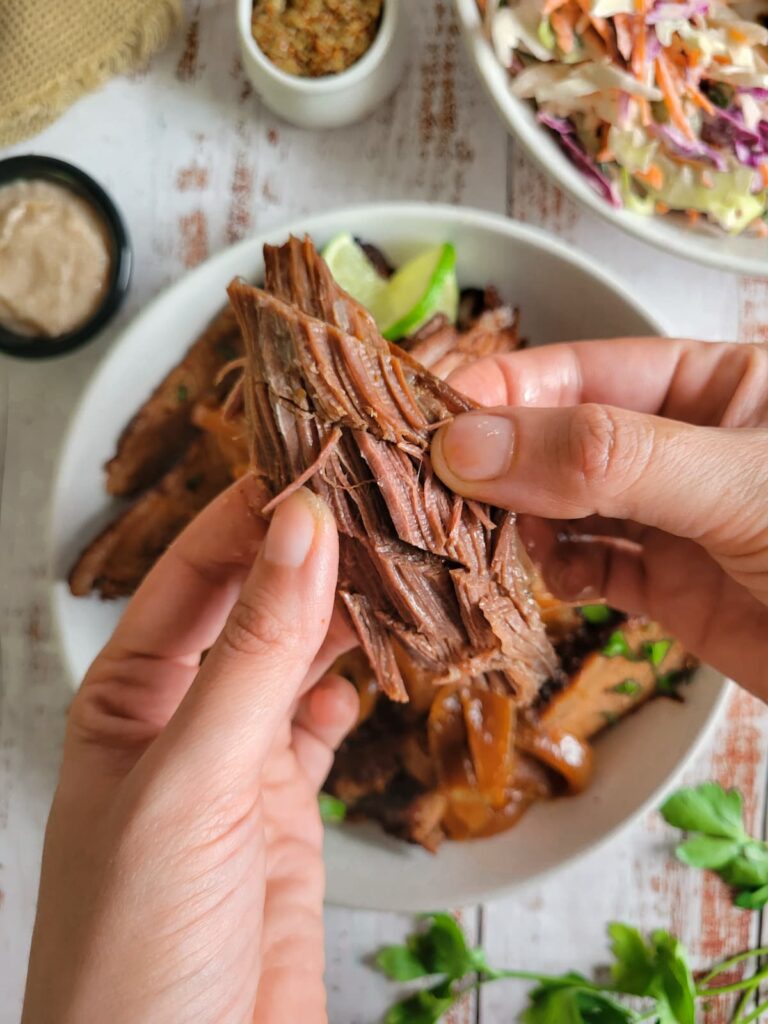 hands pulling apart some brisket over a bowl with the rest. Sauces and coleslaw in the background