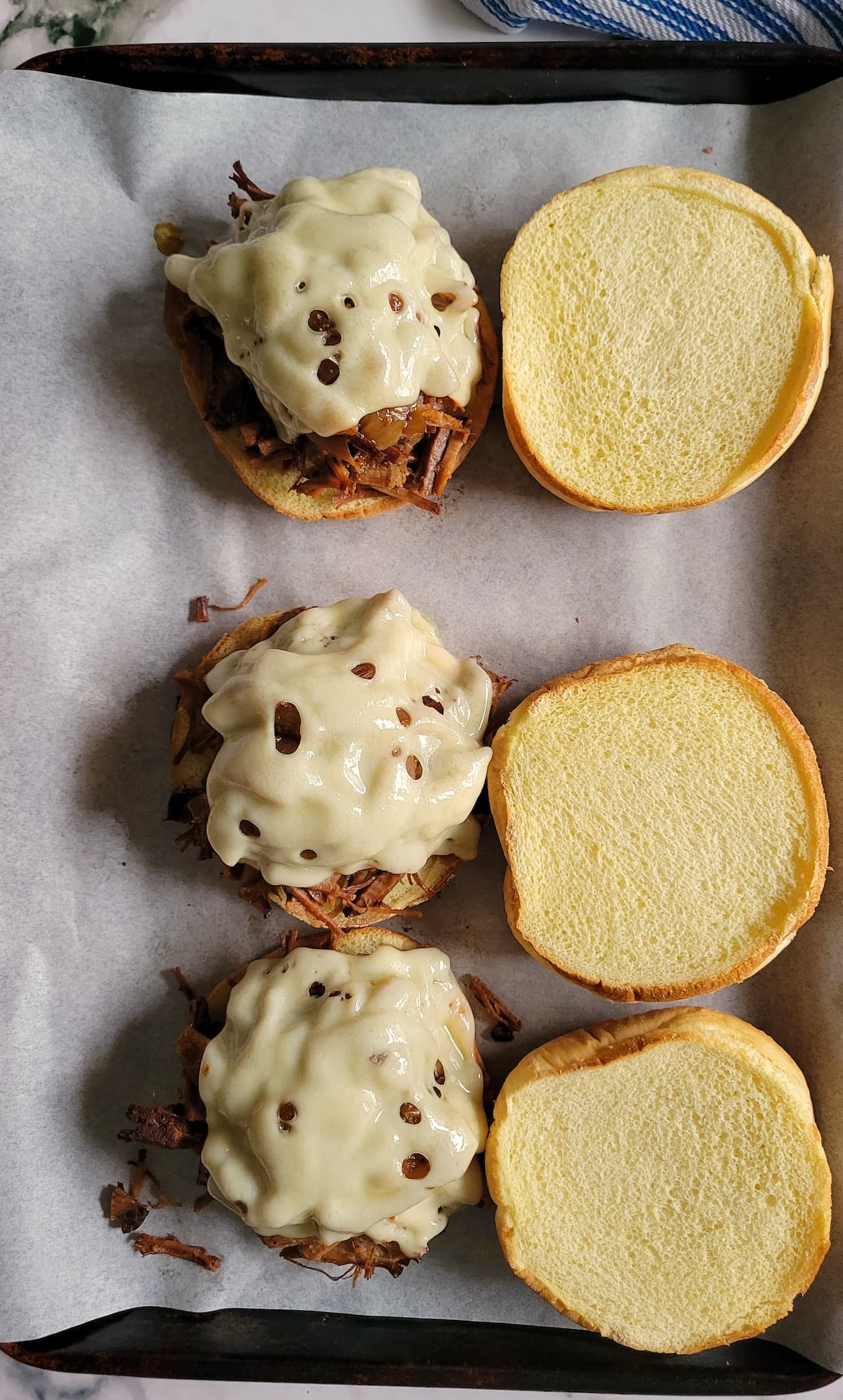 3 bottom buns loaded with brisket and cheese next to a top bun on a parchment lined baking sheet