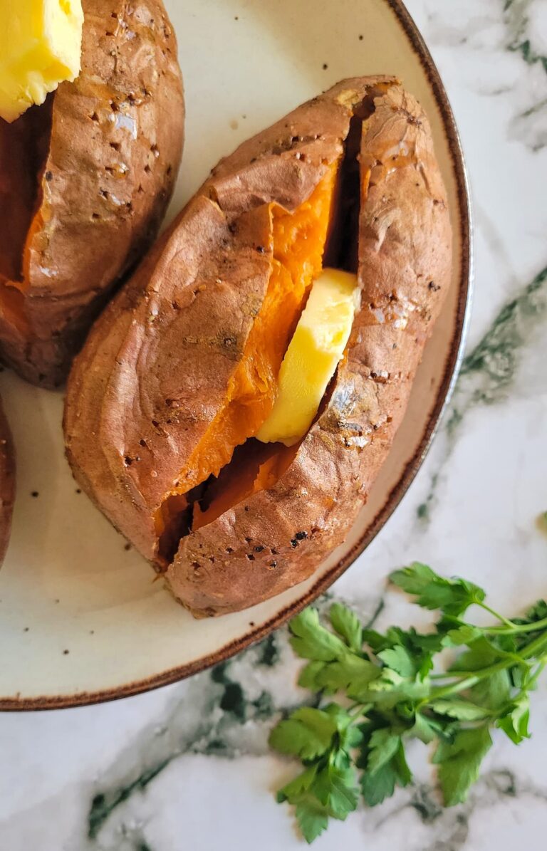 baked sweet potato cut open with a slice of butter, herbs in the background