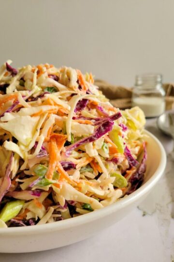 side view of a bowl of coleslaw, spoons and creamy dressing in the background