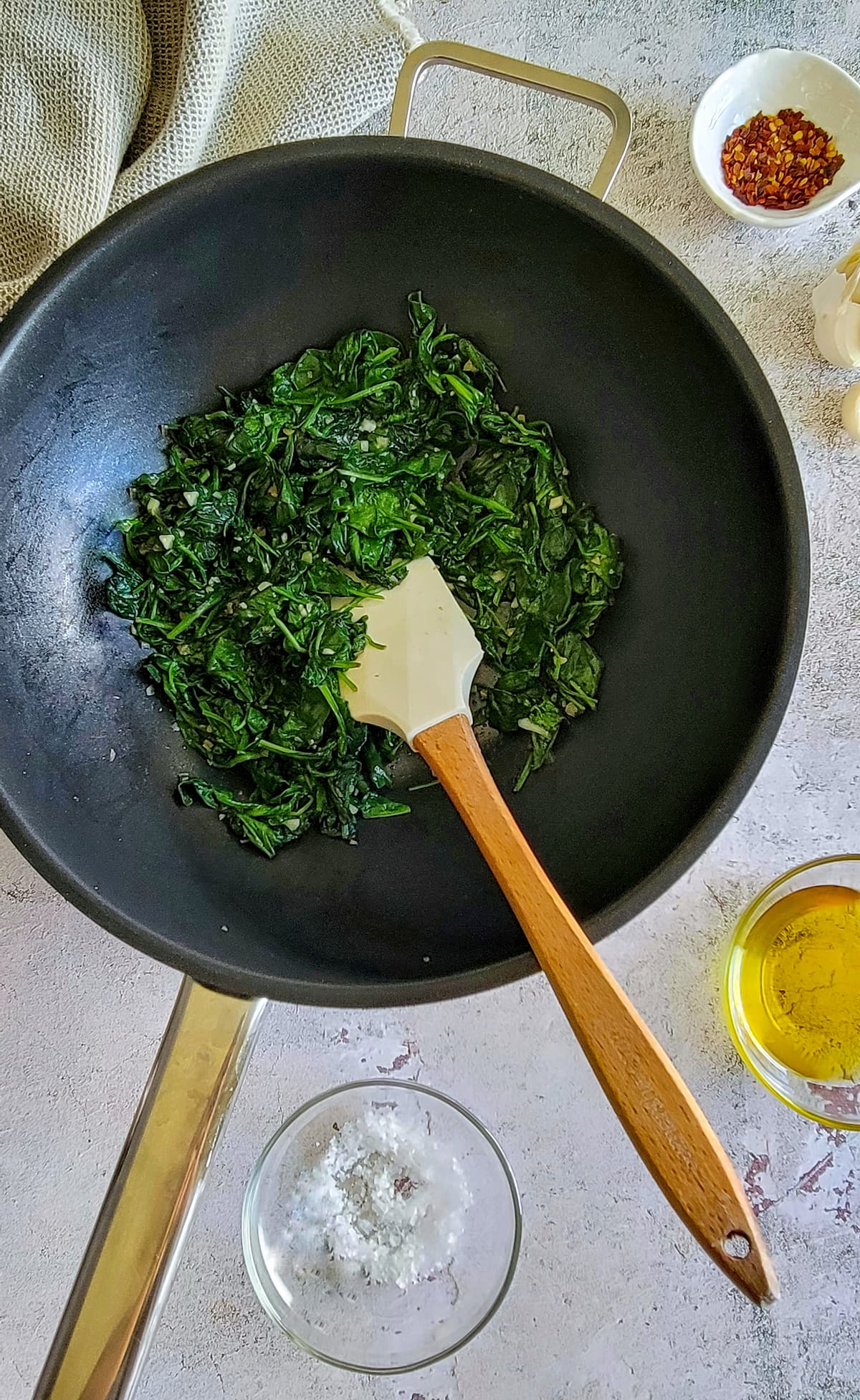 spinach in a wok with a spatula, chili flakes, olive oil and salt in the background
