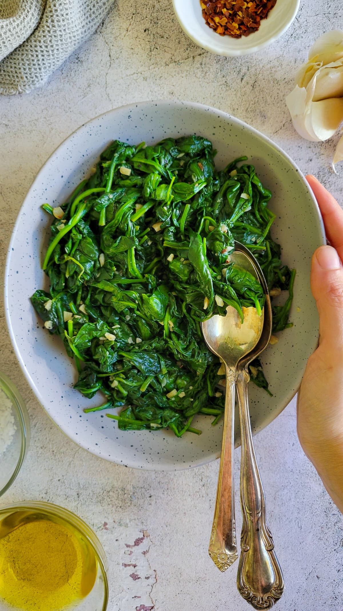 hand holding a bowl with cooked spinach and minced garlic, two spoons in the bowl, chili flakes, garlic cloves and olive oil around it